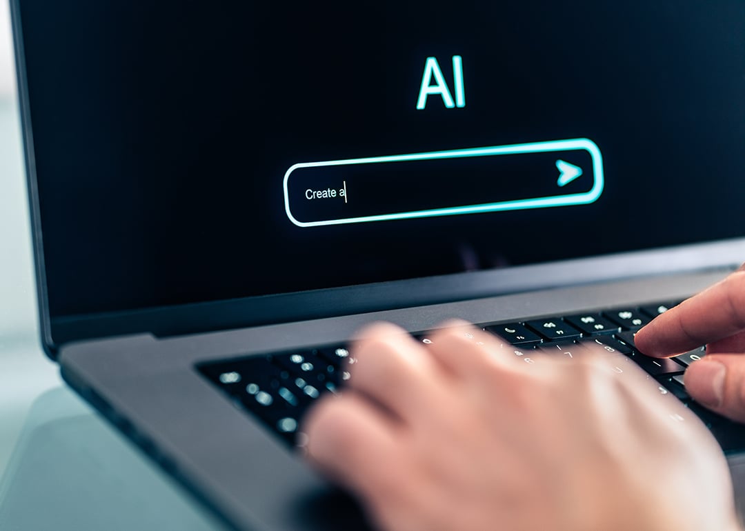 A close-up shot shows a person's hands typing on a laptop keyboard, with the screen displaying the word "AI" above a search bar that has text input. This image visually represents the interaction between humans and artificial intelligence, possibly in the context of searching for information, developing AI, or using AI-powered tools.
